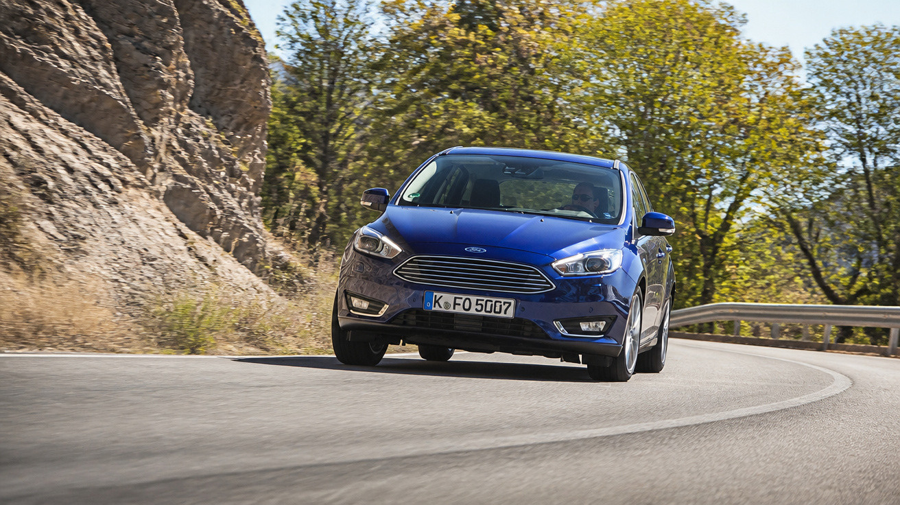 Blue Ford car driving on a curved mountain road with trees and rocky cliffs in the background.