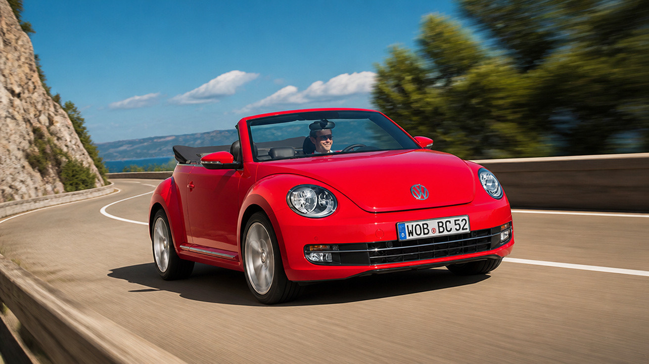 Red Volkswagen Beetle convertible driving on a coastal road with a driver wearing sunglasses.