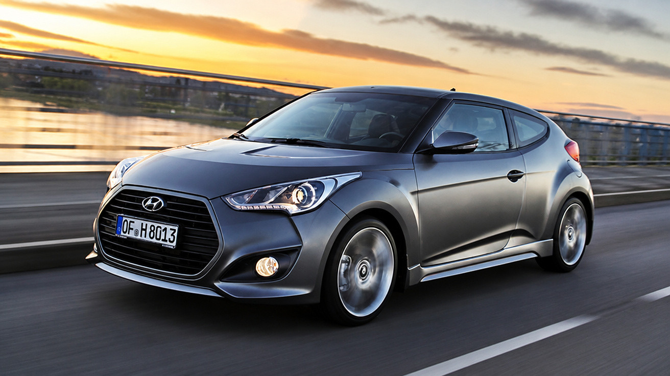 Grey Hyundai hatchback car driving on a road beside a river during sunset.