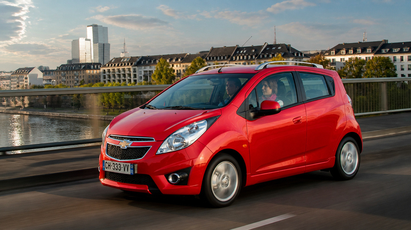 Red Chevrolet compact car driving on a bridge over a river with buildings in the background.