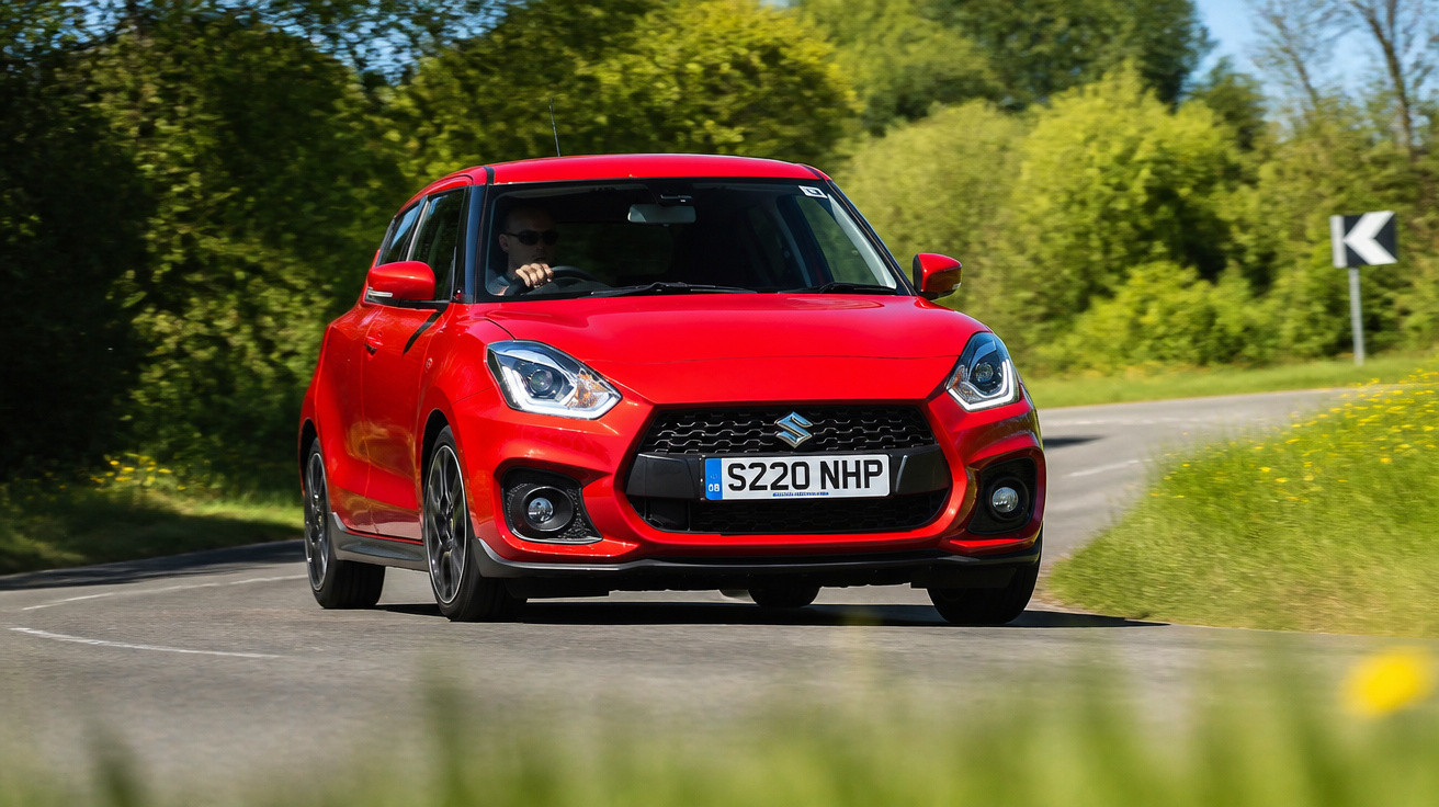 Red Suzuki Swift car driving on a curvy road with greenery and a chevron road sign in the background.