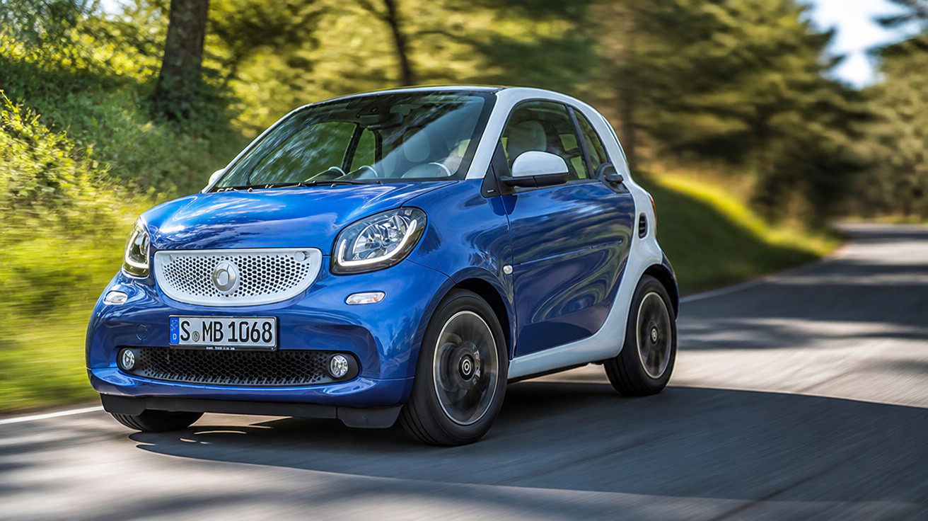Blue and white Smart car driving on a sunlit country road with trees and greenery in the background.