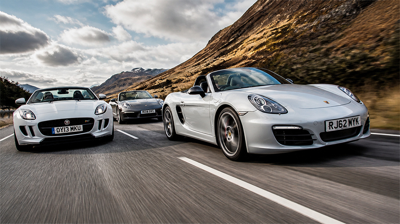 Three sports cars driving on a mountain road under a partly cloudy sky in a dynamic action shot.