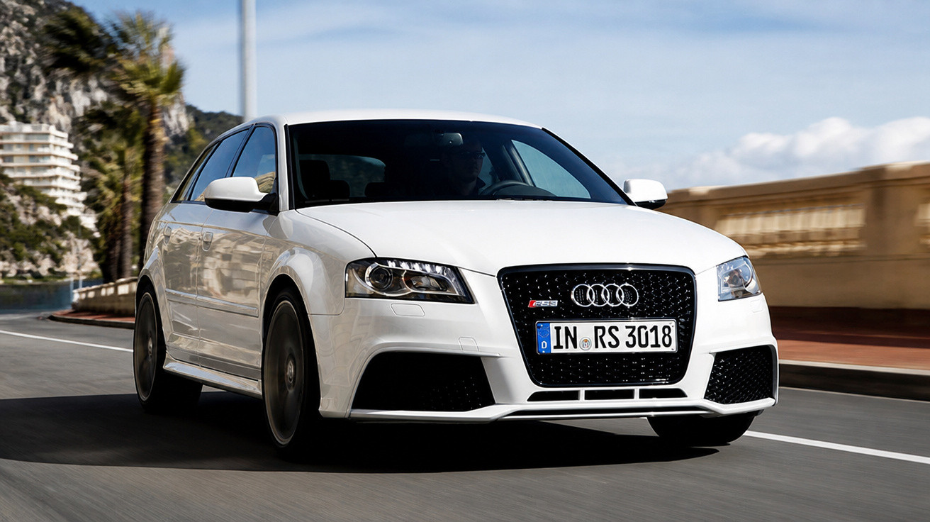 White Audi RS3 driving on a coastal road with palm trees and cliffs in the background.
