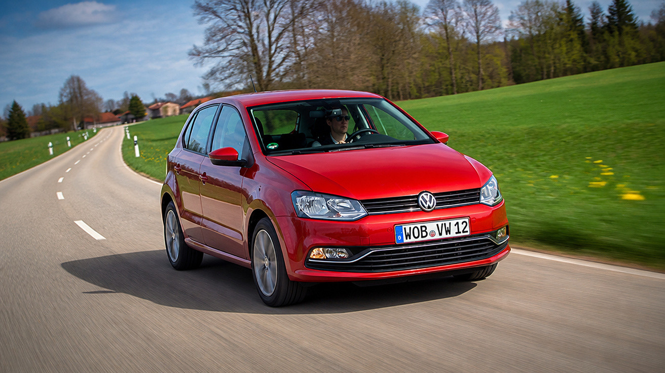 Red Volkswagen hatchback driving on a rural road with green fields and trees in the background.