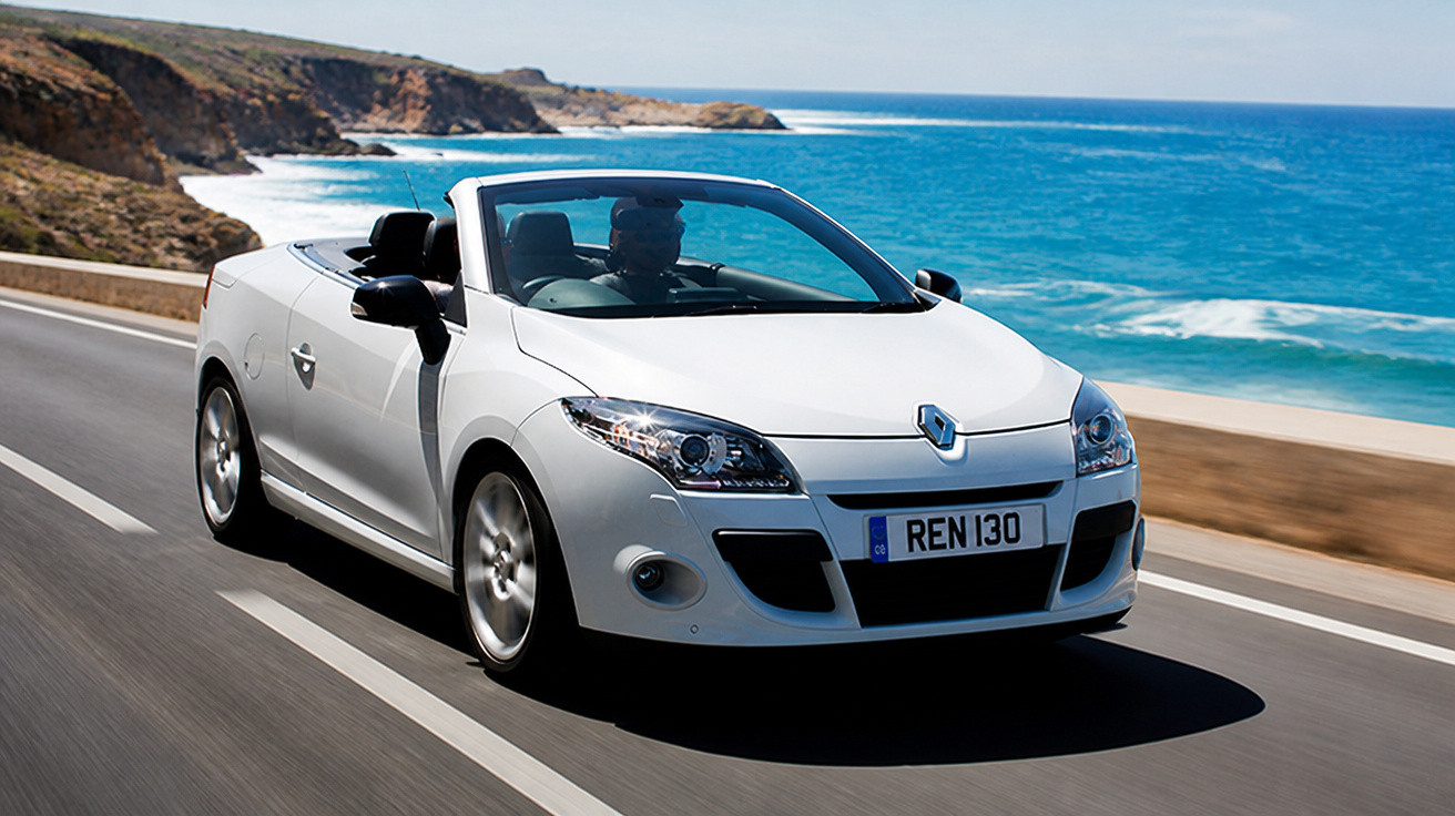 White Renault convertible car driving along coastal road with blue sea and cliffs in background.