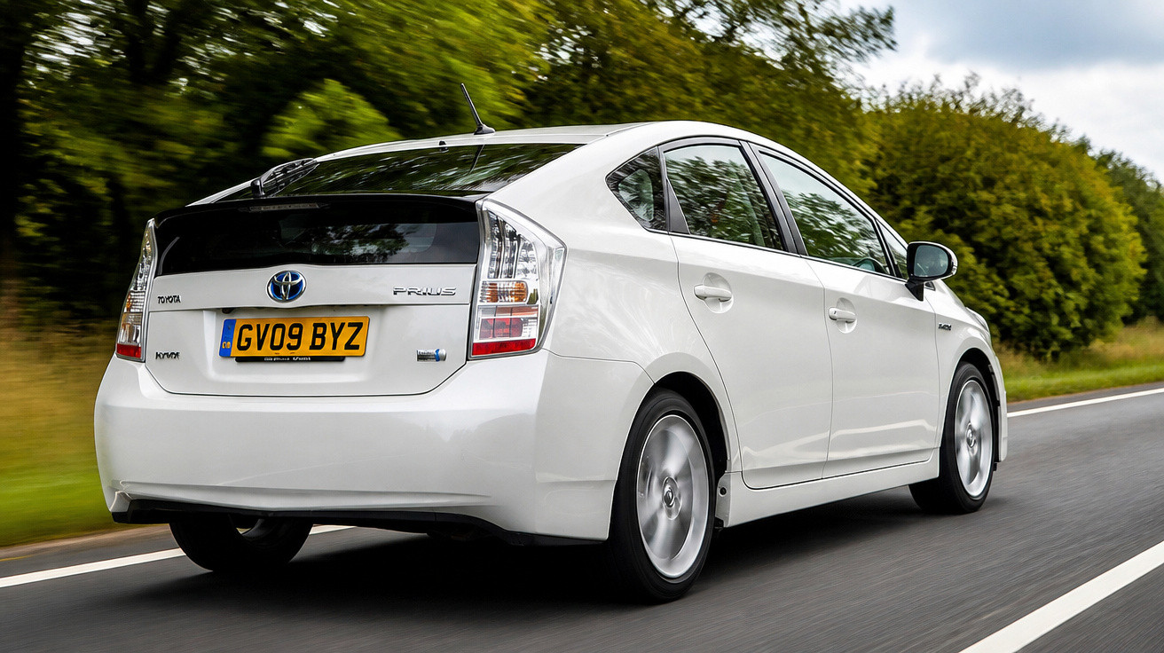 White Toyota Prius hybrid car driving on a rural road with greenery and trees in the background.
